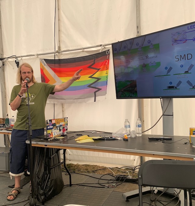 Arne standing in front of a pride flag, with a large flat screen TV to his left, showing helpful guidance for those new to soldering..."
